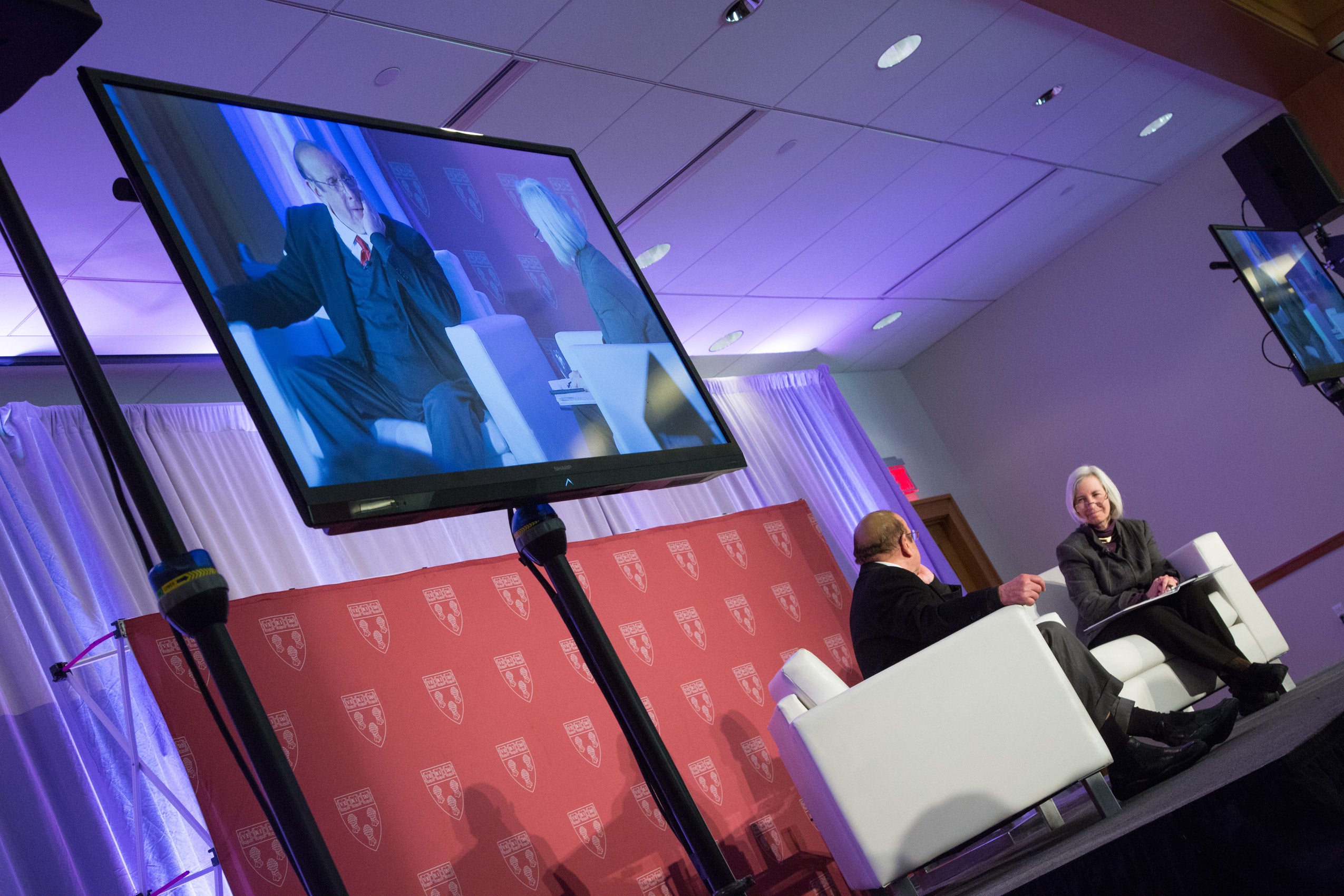A man and a woman seated on stage having a conversation with a large screen next to them