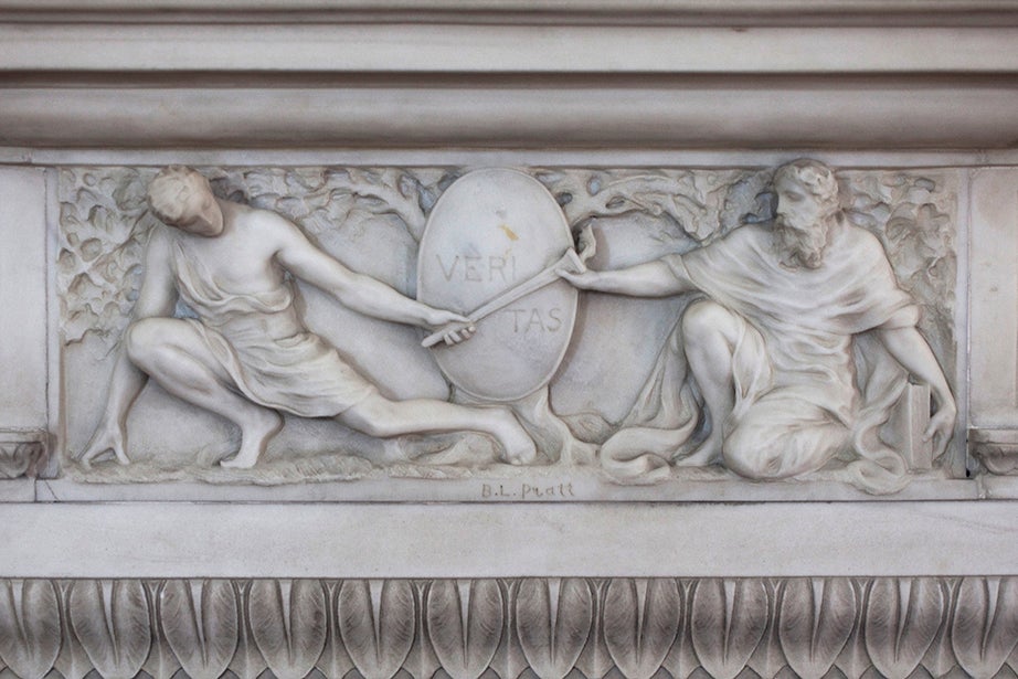 A closeup of white carved marble in the shape of two men wearing robes, one is passing a scroll to the other