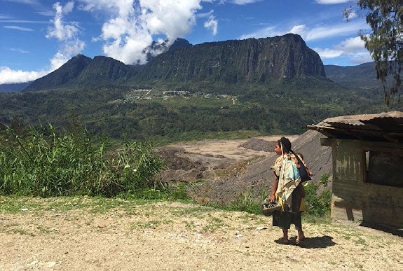 A native woman standing beside a hut with a large mountain in the background