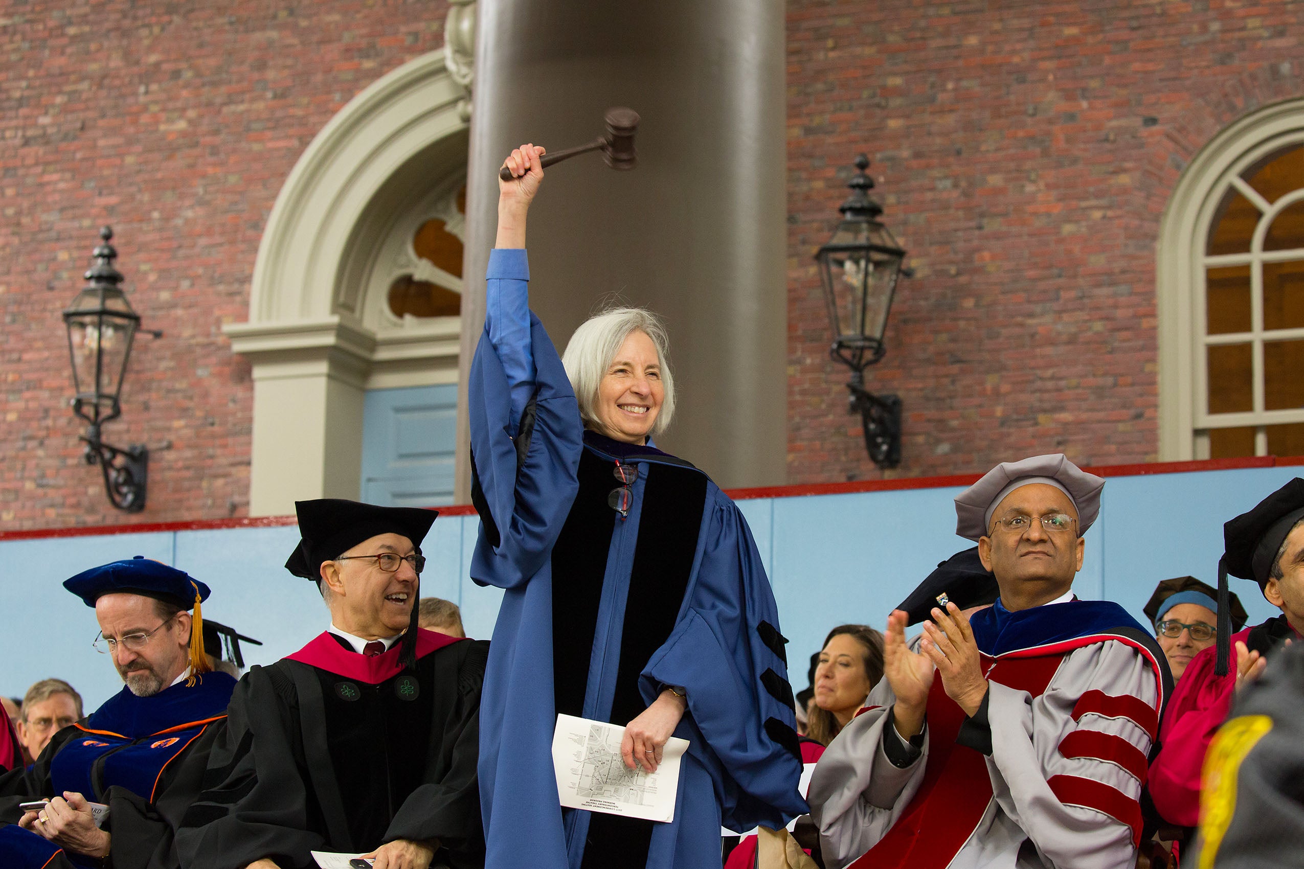 A woman in cap and gown, standing and smiling with a gavel raised into the air