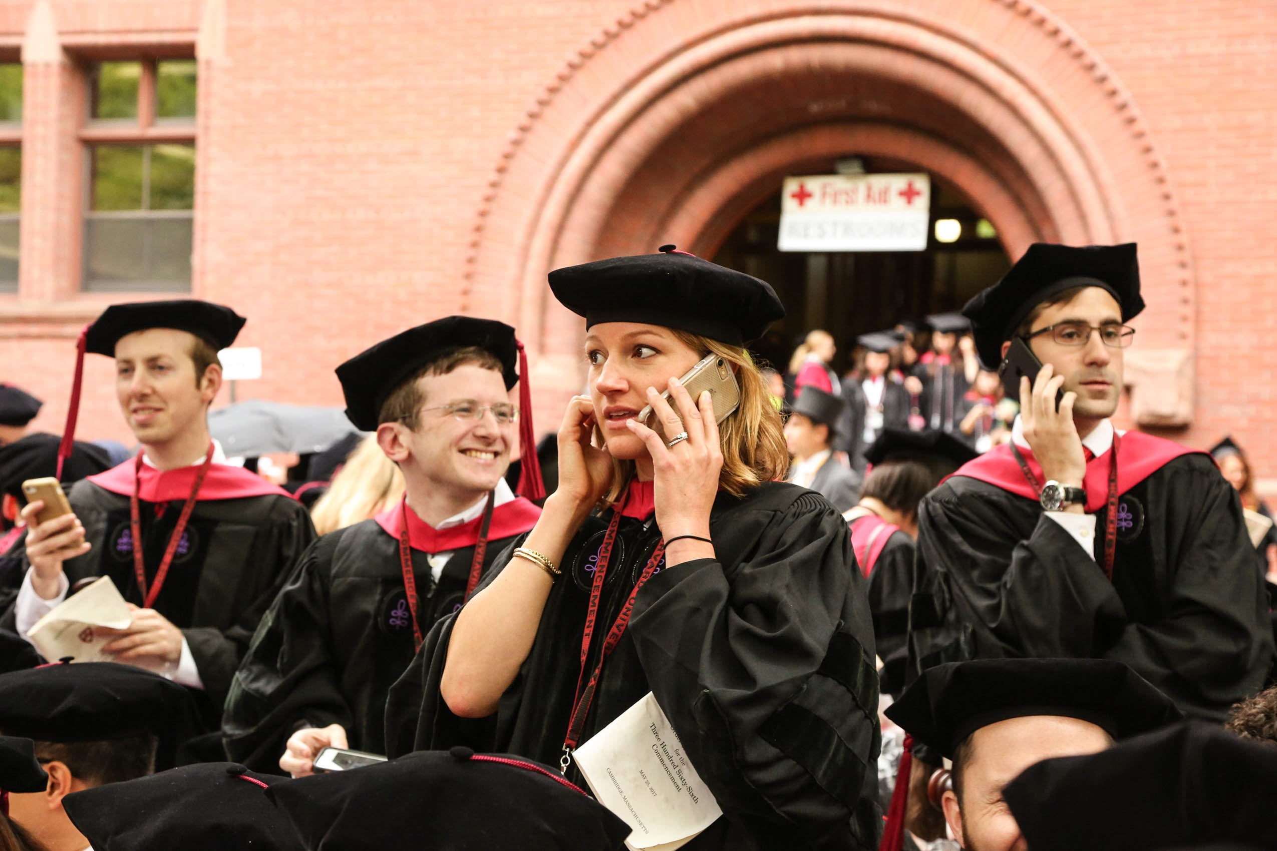 Students in caps and gowns, seated on Holmes Field. Two are speaking on their phones