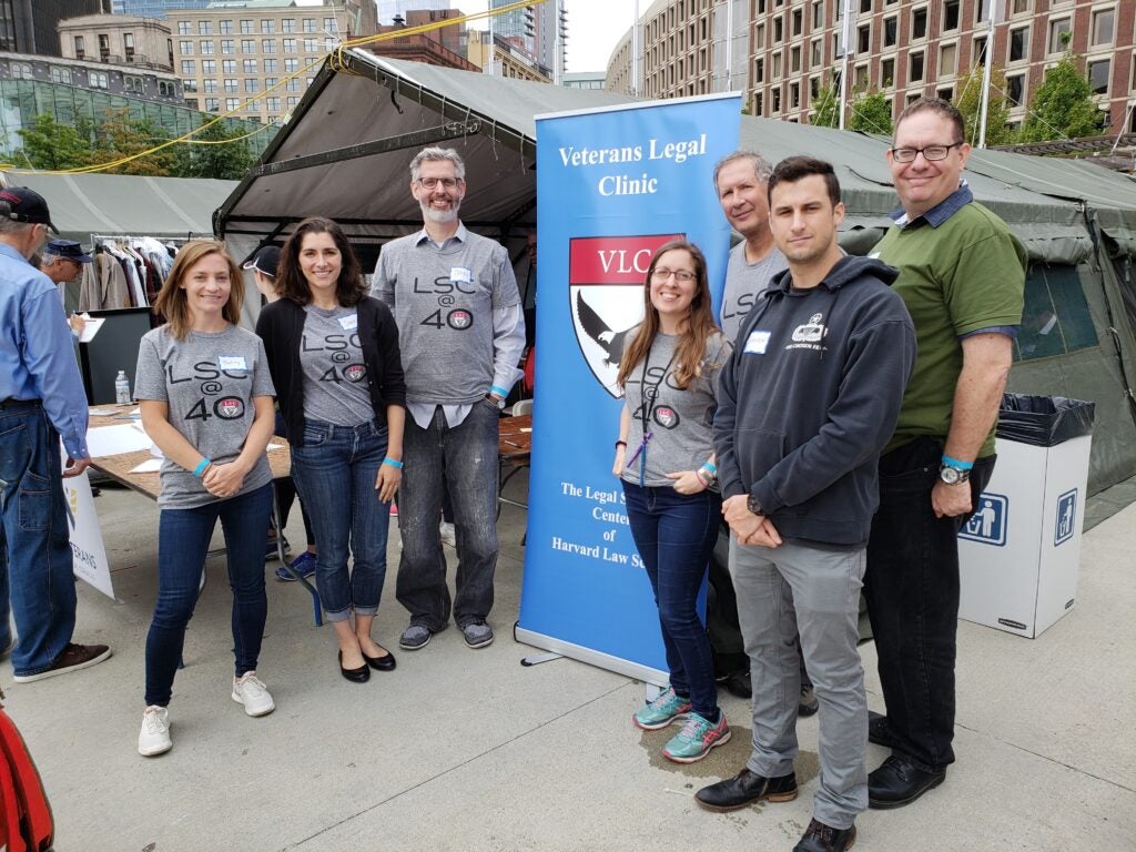 Legal Services Center stand in front of a large olive tent and a blue Veterans Legal Clinic banner with the VLC shield on it. From left: Betsy Gwin, Dana Montalto, Dan Nagin, Julia Schutt, Keith Fogg, clinical student Steven Kerns, Evan Seamone