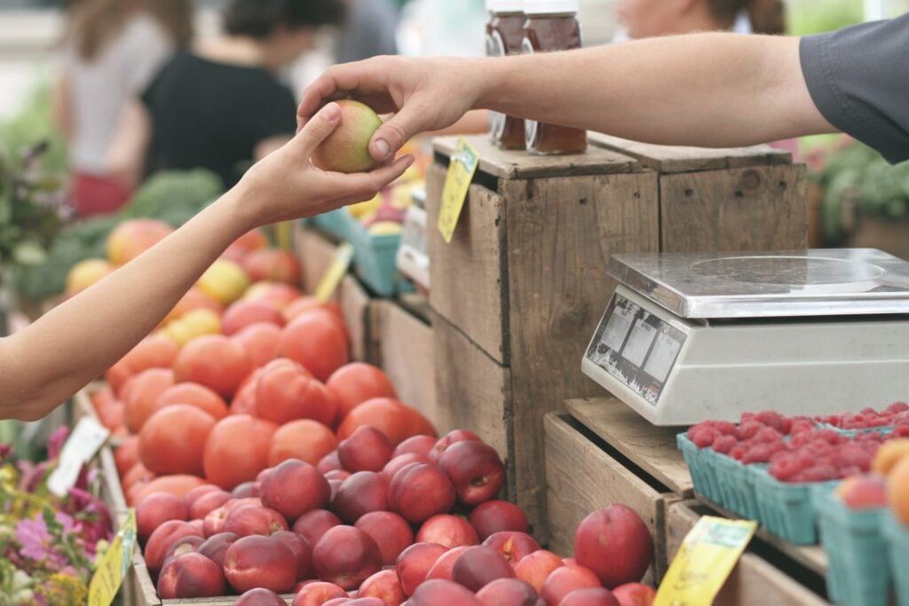 A person passes an apple to a customer at a market over containers of fruit