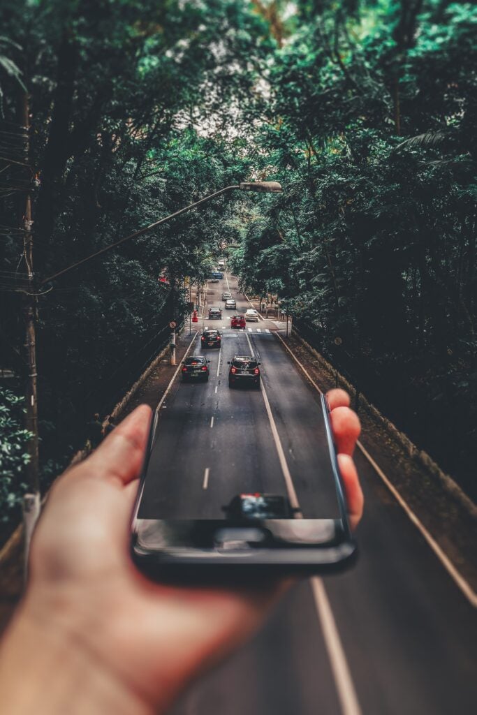 Hand holding a smartphone that merges into the background which shows a road lined with trees and cars driving away
