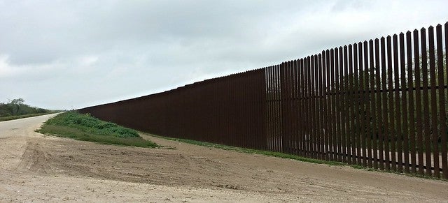 The U.S.-Mexico border shows a tall brown fence along a side of road