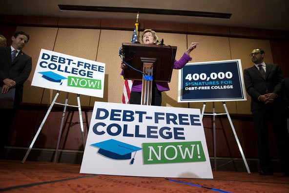 Elizabeth Warren stands at a podium with signs to her left, right, and front reading 'debt-free college now' and '400,000 signatures for debt-free college'. Security detail stands to her left and her right.