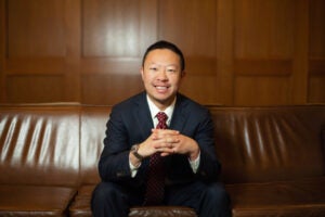 Photo of Alex Chen sitting on a brown couch in front of a wood paneled wall, fingers laced in front of him.
