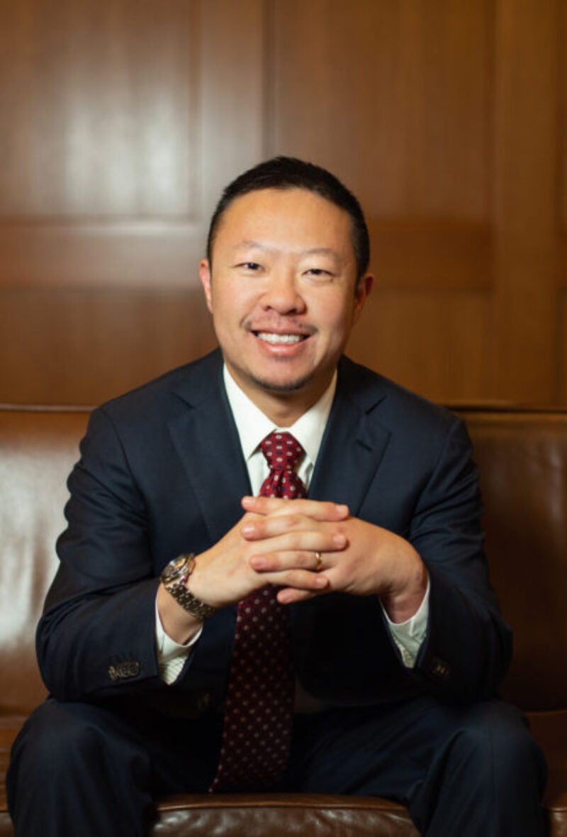 Photo of Alex Chen sitting on a brown couch in front of a wood paneled wall, fingers laced in front of him.