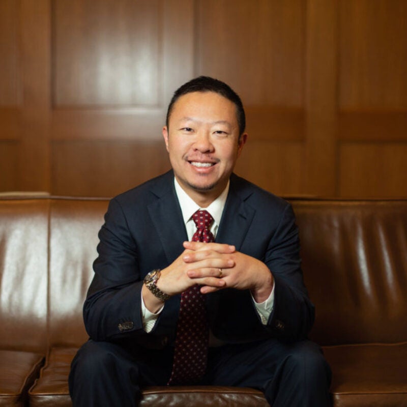 Photo of Alex Chen sitting on a brown couch in front of a wood paneled wall, fingers laced in front of him.