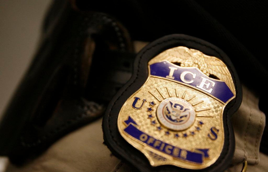 An ICE officer badge in gold and blue is shown clippped to a belt of an officer