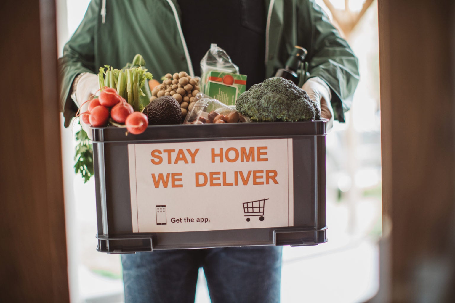 man in green shirt holds a box of produce that reads "stay home we deliver"