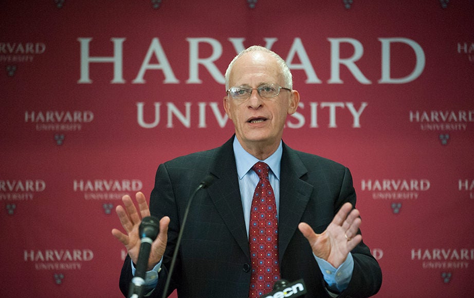 Oliver Hart speaking at a podium with microphones, with a Harvard backdrop behind him
