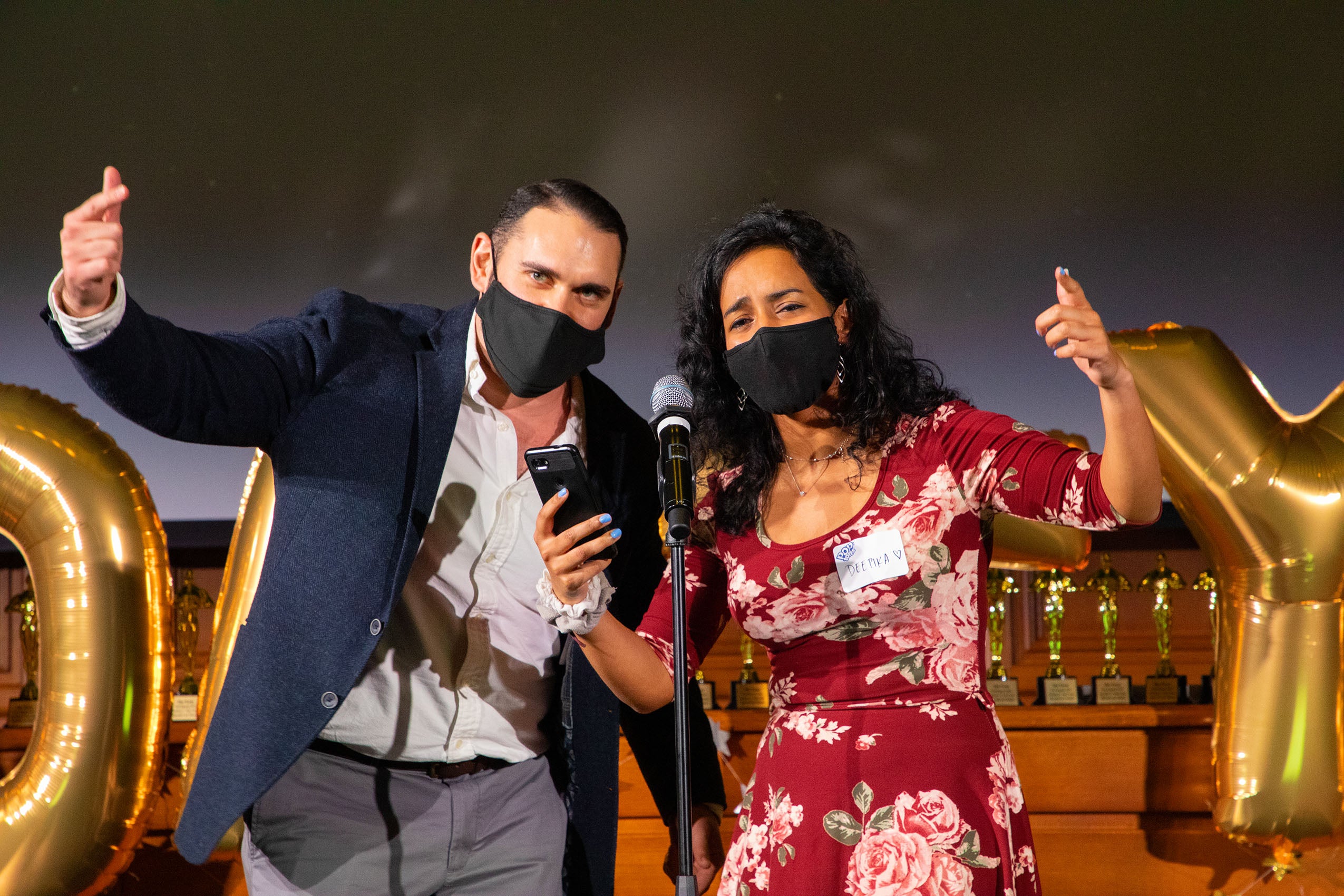 Man and woman wearing face masks, standing on stage at a microphone, looking at a cellular phone and gesturing.