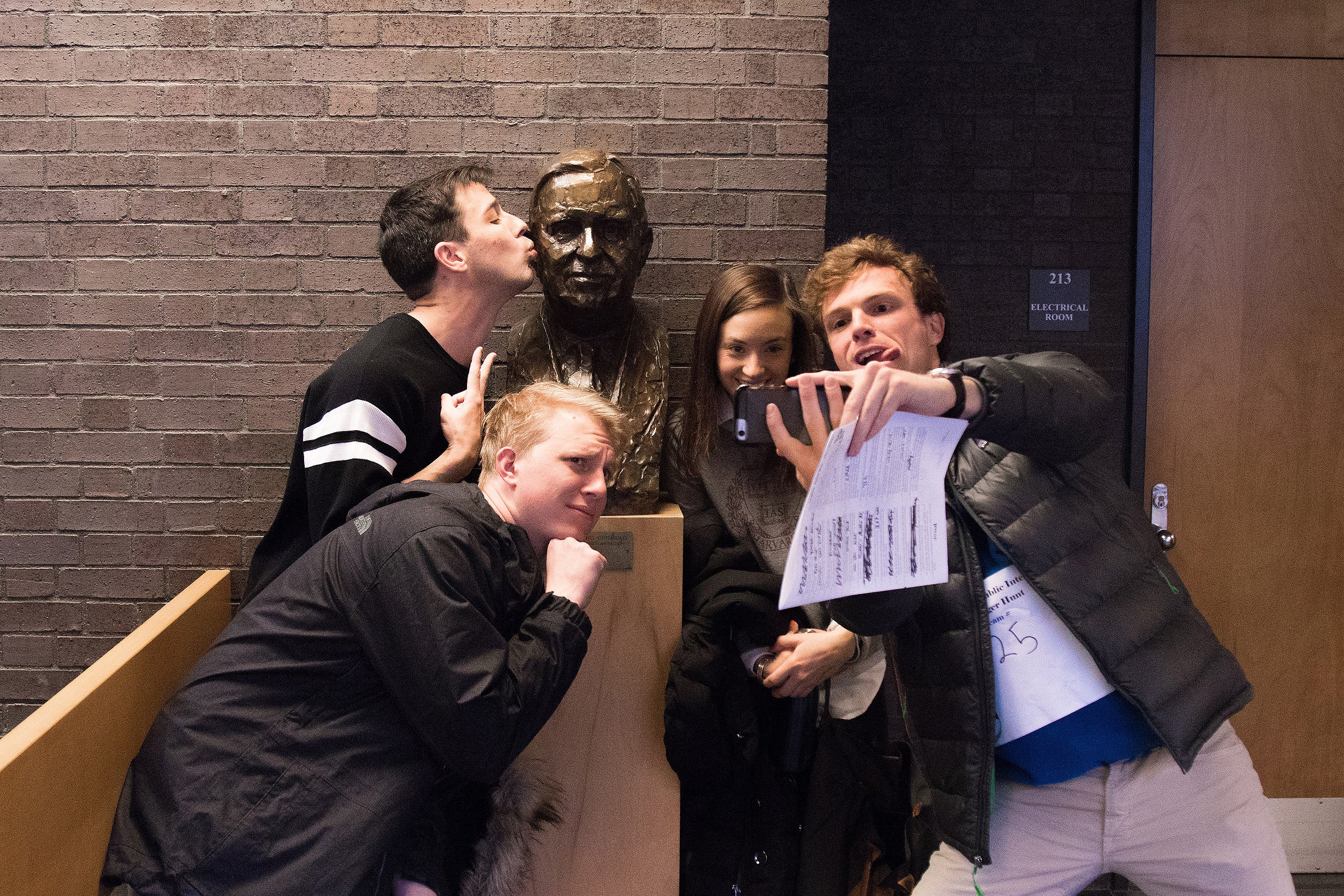 Four students posing in front of a bust, one of them kissing it on it's cheek