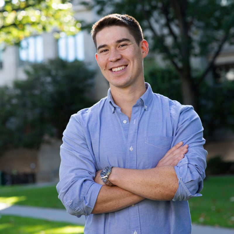 A man in a blue shirt stands outside in front of a building on the Harvard Law School campus.