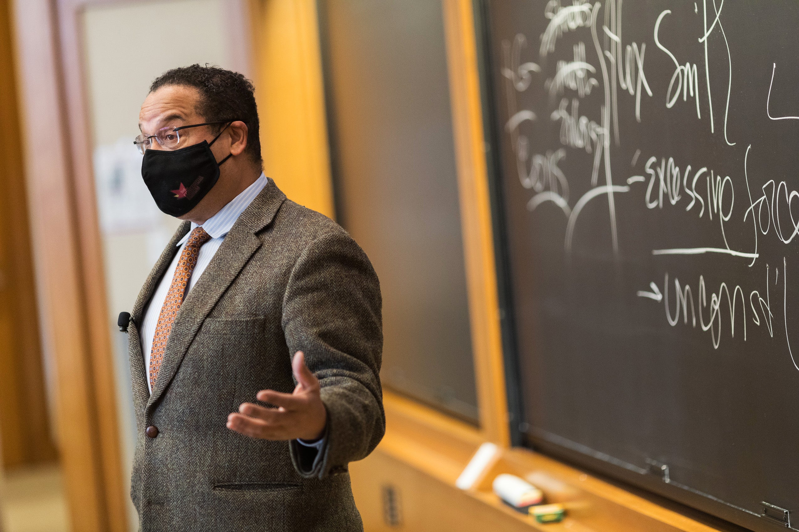 Minnesota Attorney General Keith Ellison, wearing a face mask, speaks in front of a blackboard with writing on it.