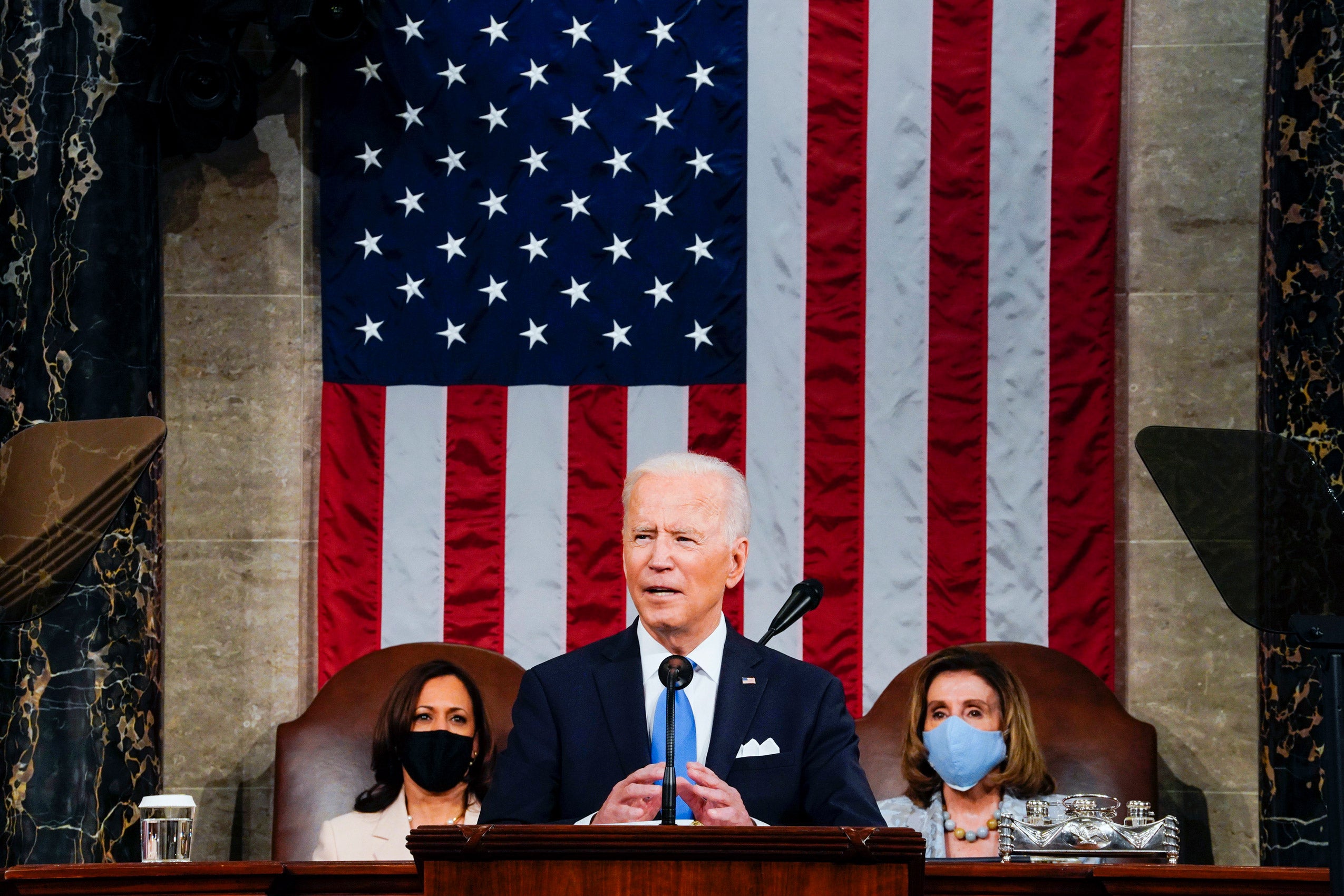 American flag on the wall in the background; President Joe Biden at a podium with Vice President Kamala Harris and House Speaker Nancy Pelosi sitting behind him.