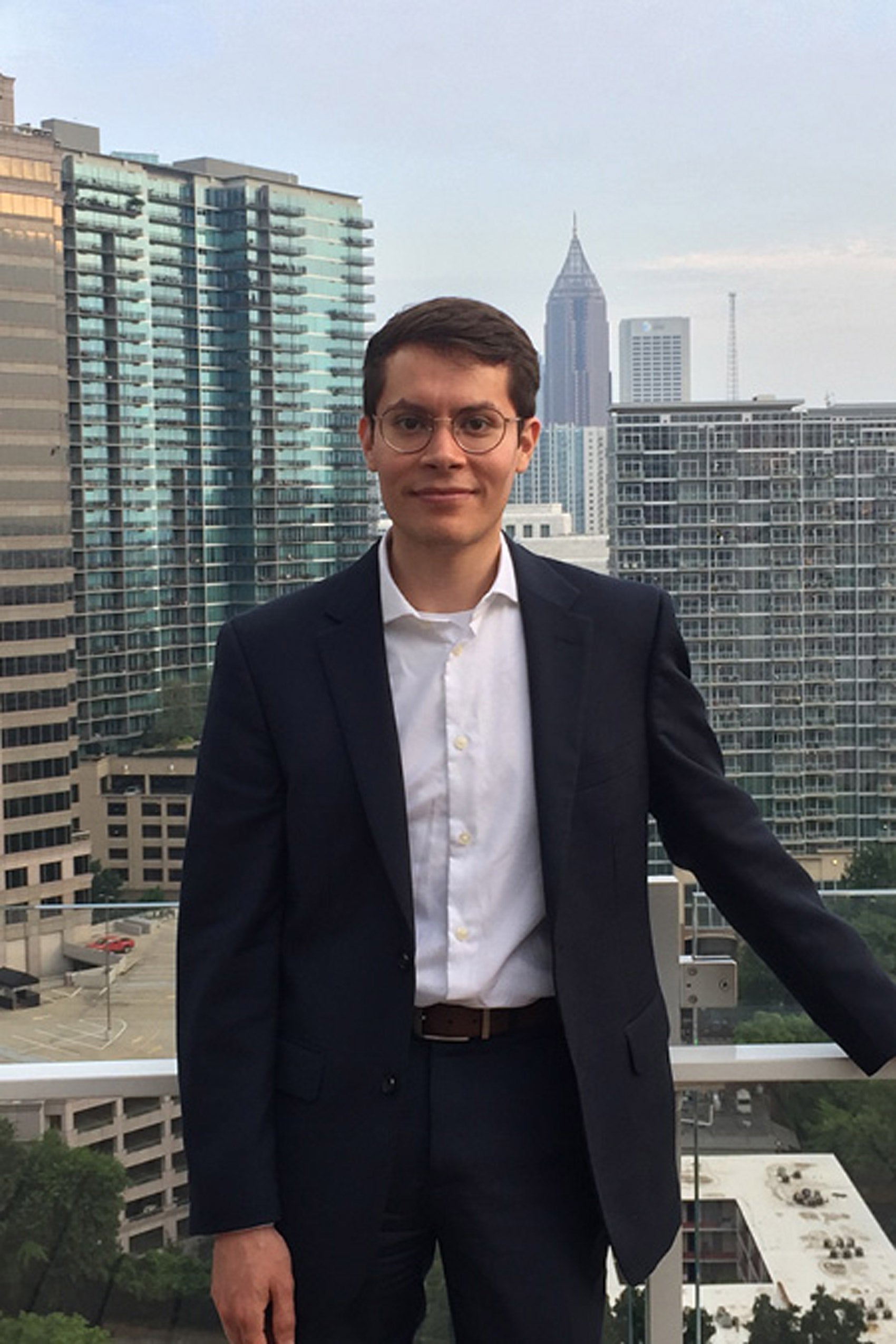 Man with brown hair and glasses, blue suit and white shirt standing in front of a window