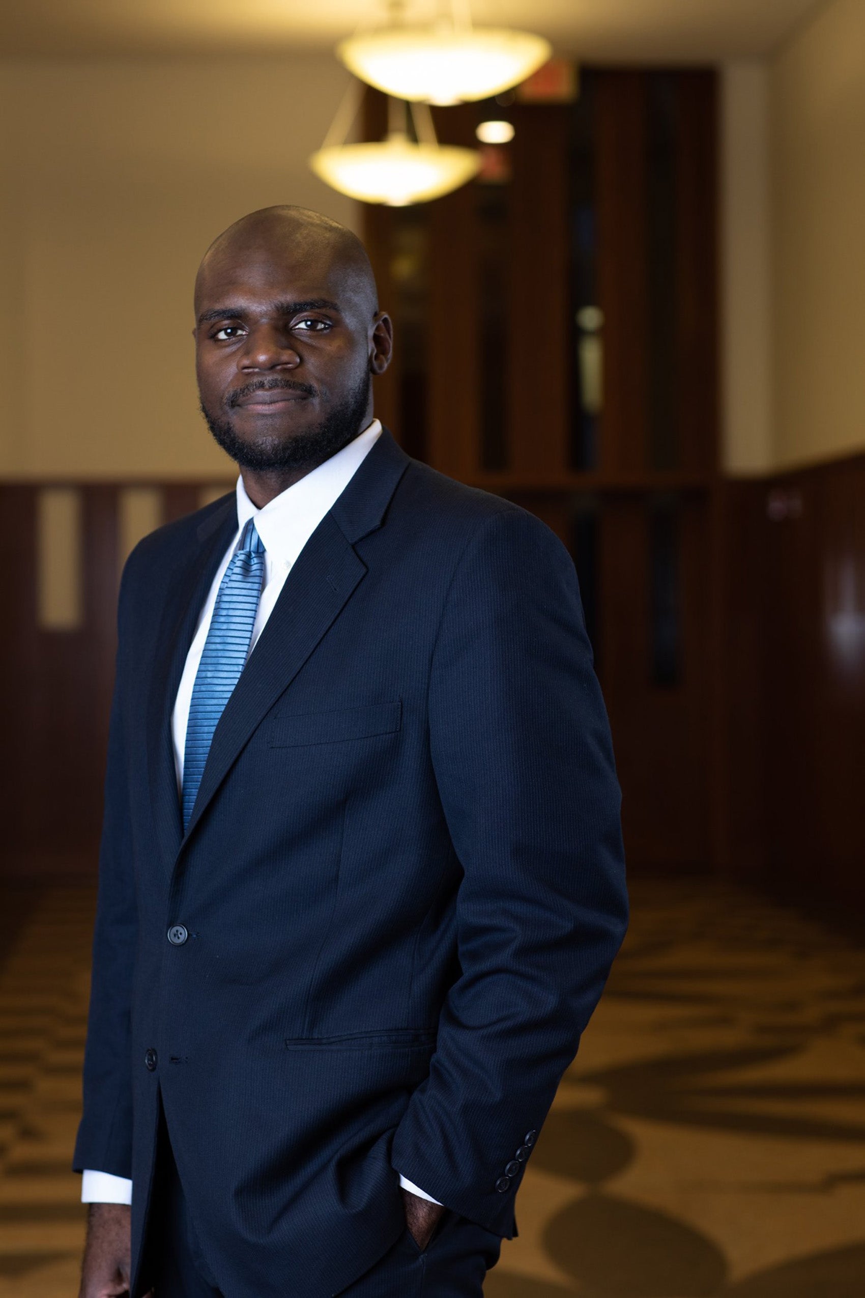 Man standing in a room wearing a navy blue suite and blue tie