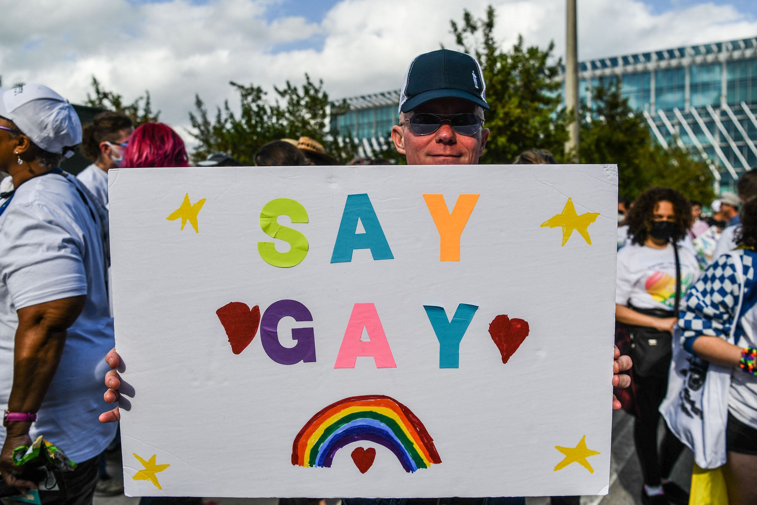 A man stands outside at a protest holding a sign that reads: SAY GAY.