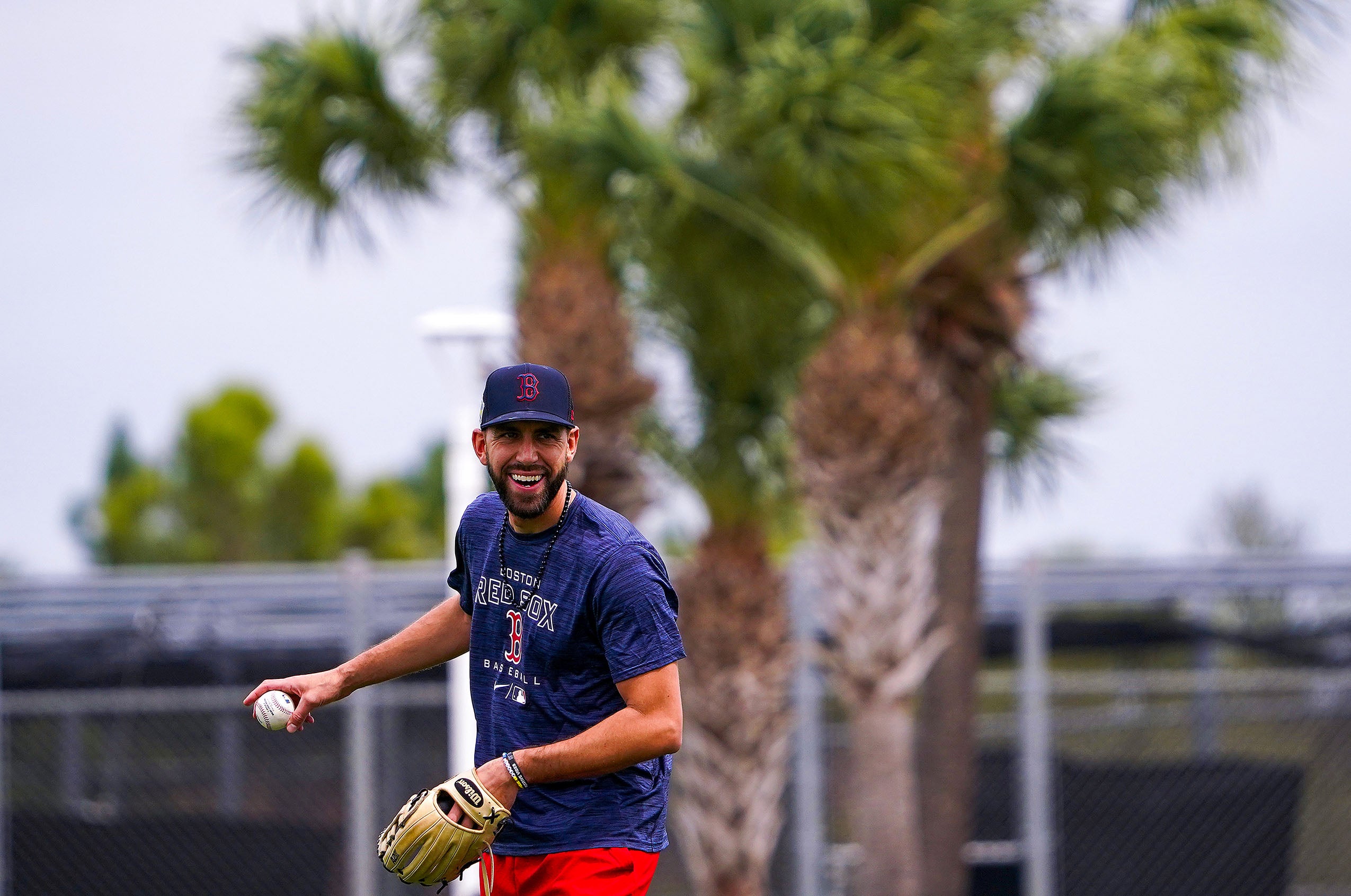 Man smiling, holding a baseball and baseball glove, wearing a Boston Red Sox hat and t-shirt. Behind him is a palm tree and fence..