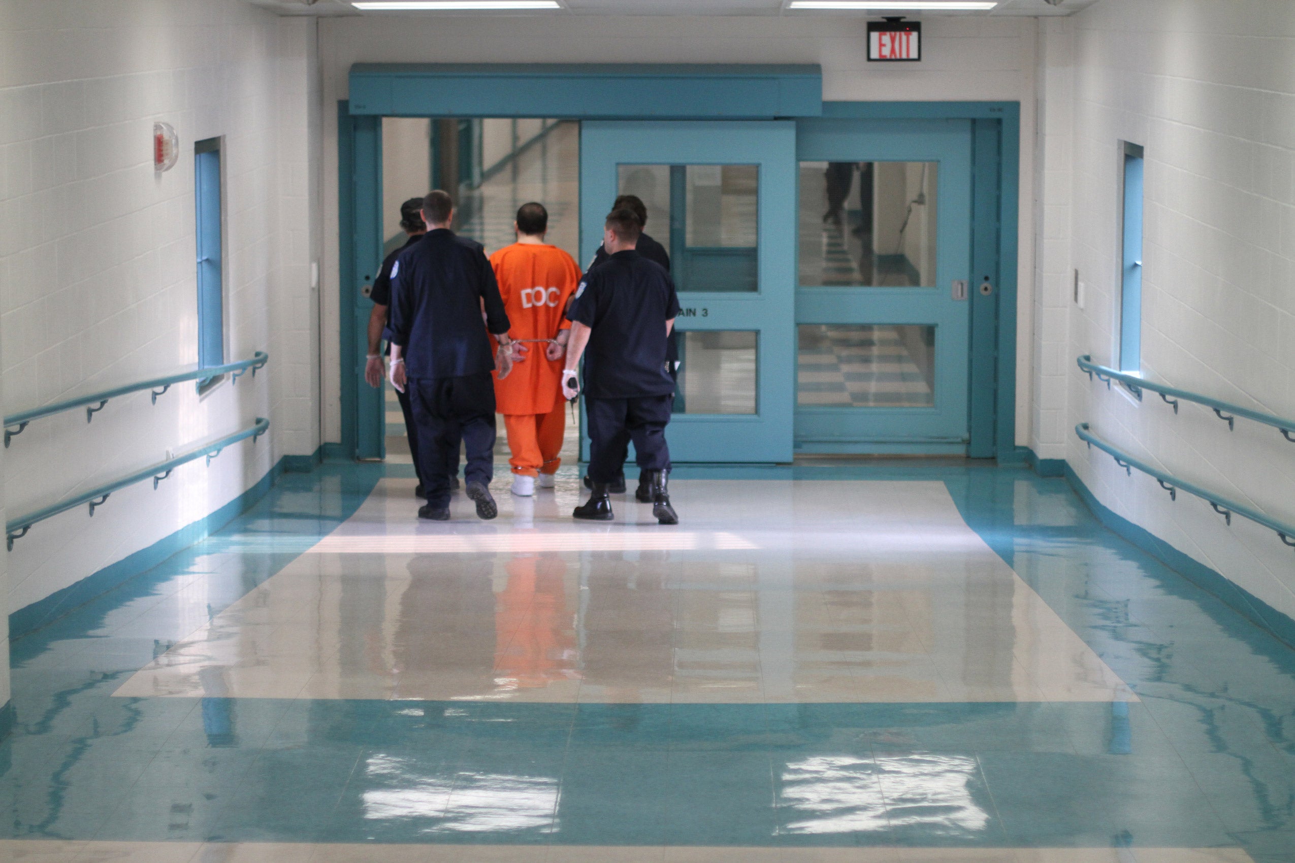 Man in orange jumpsuit wearing handcuffs is flanked by four police officers as they walk down a hallway.