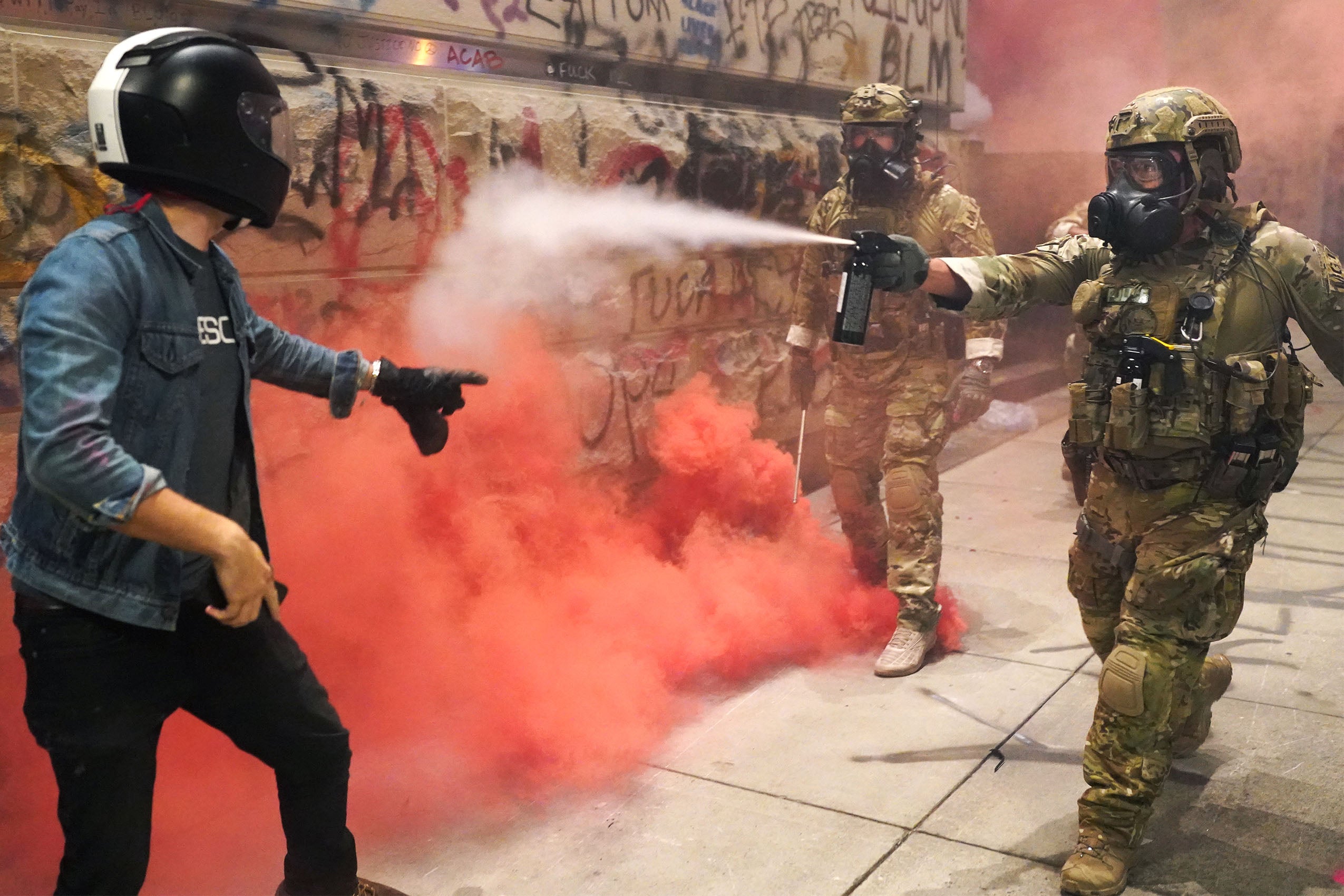 A federal officer in a camouflage uniform wearing a gas mask pepper sprays a protester wearing a motorcycle helmet next to a graffiti covered building.