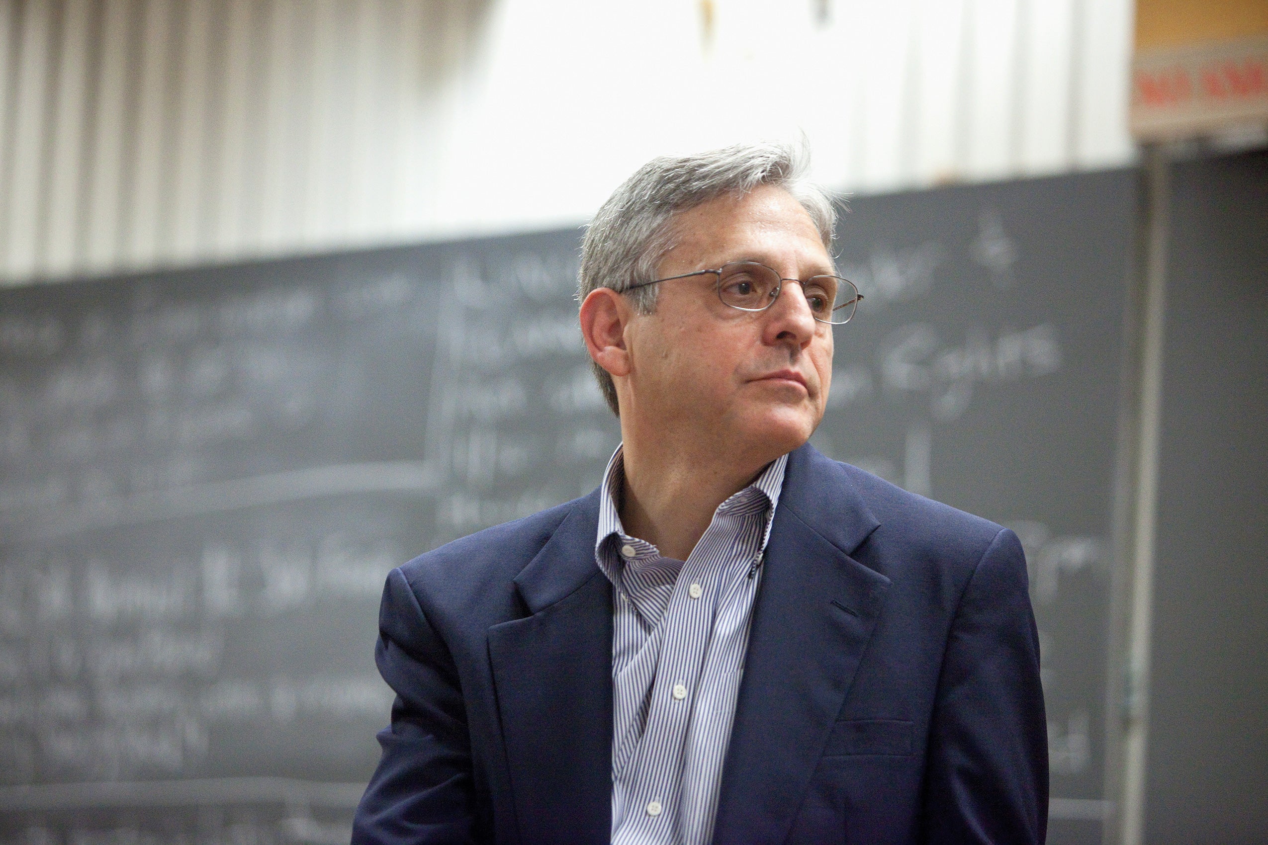 Merrick Garland looking to the right wearing a blue blazer in front of a chalk board