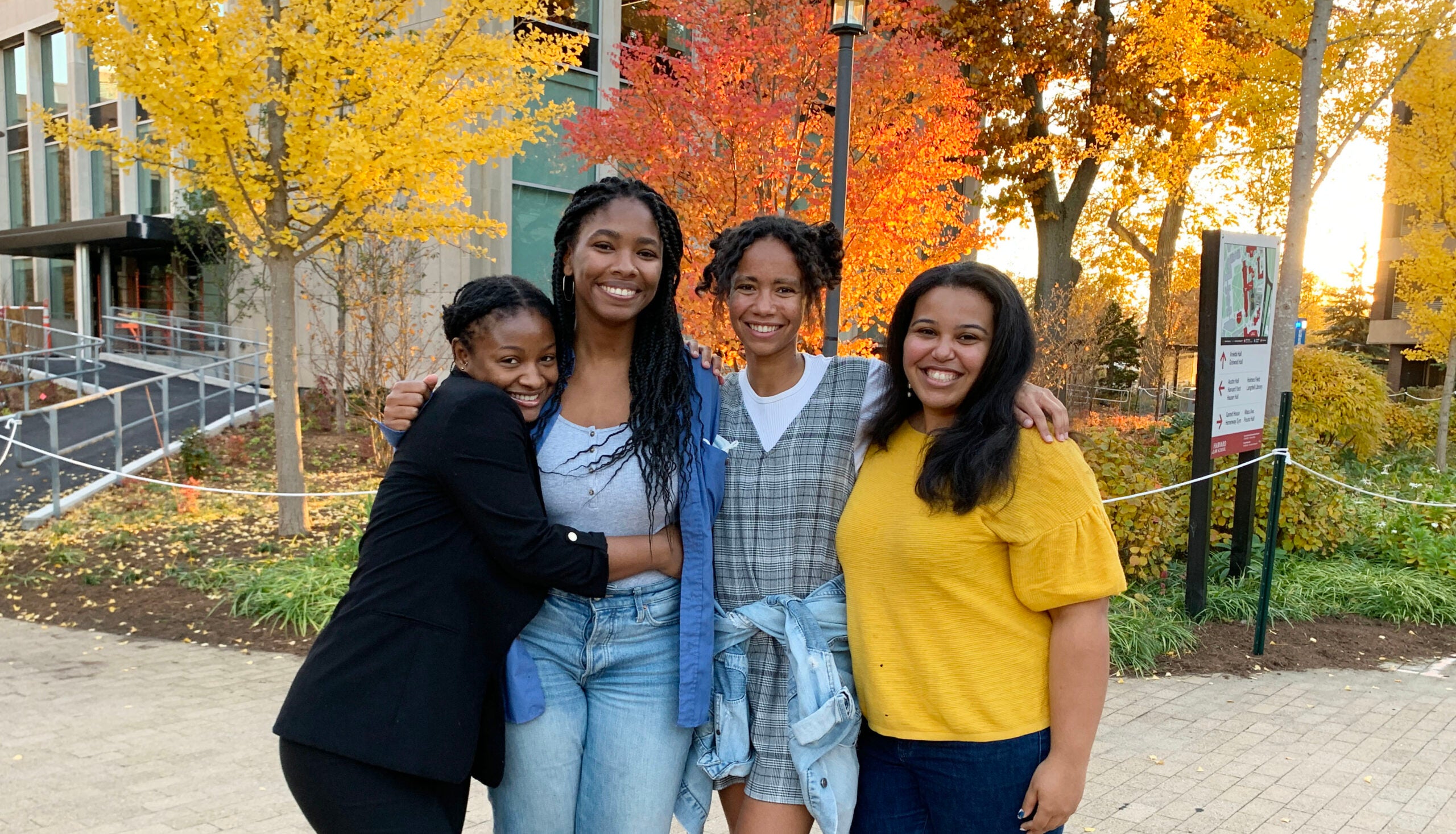 Four women standing outside, side-by-side, one woman hugging another. Trees with orange and yellow leaves behind them.