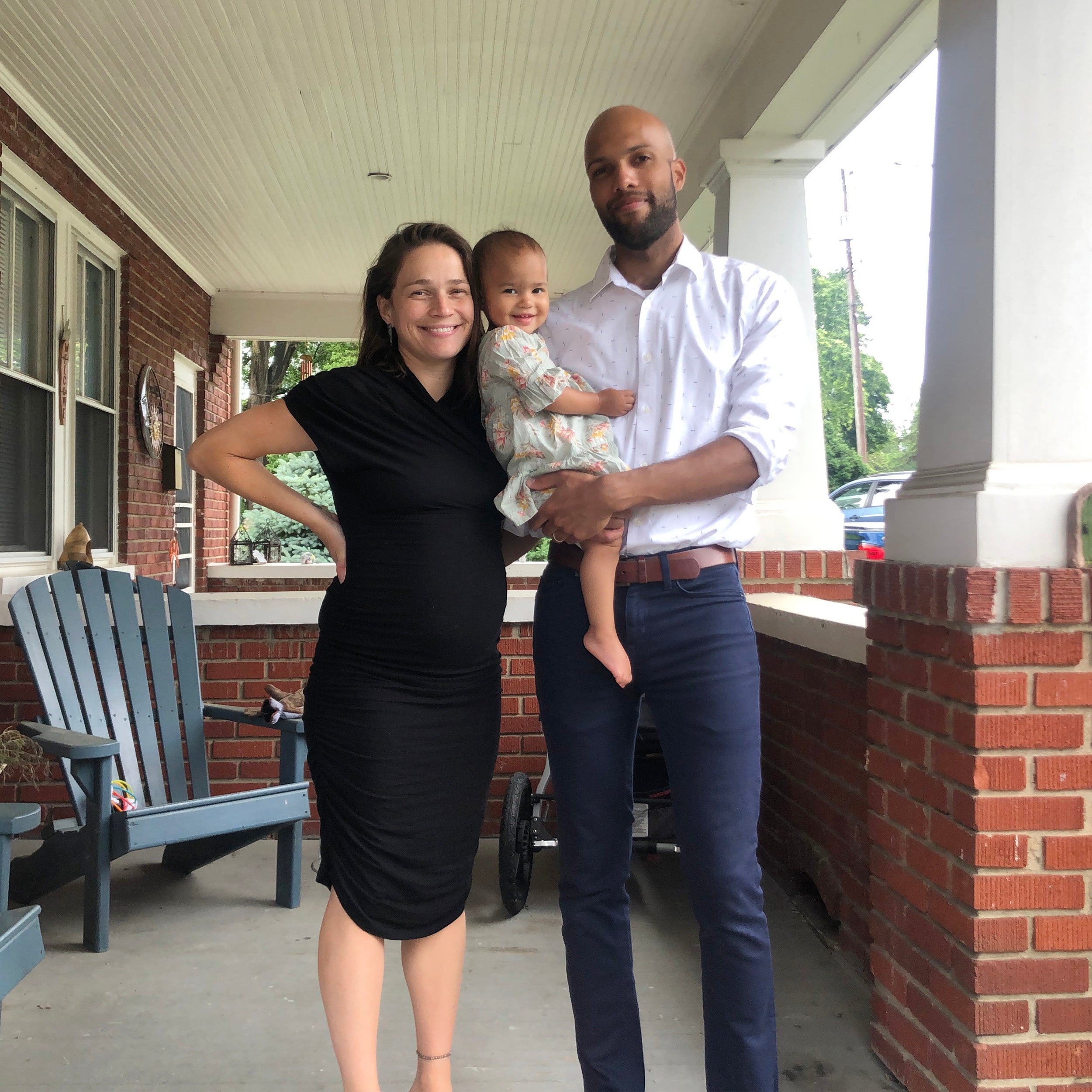 A woman in a black dress standing on a porch with a man in a white shirt holding a baby