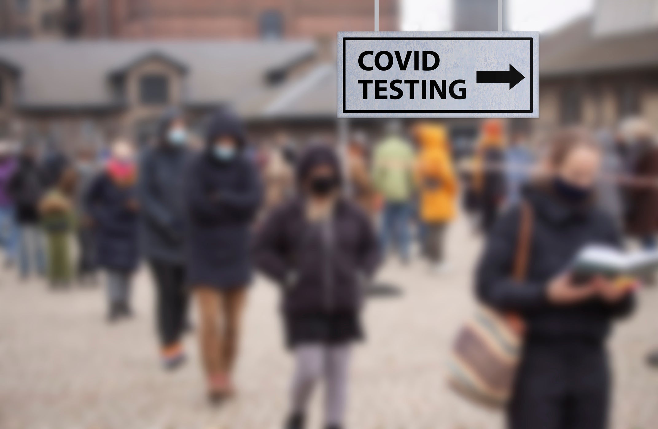 Line of people outside wearing face masks and winter coats. Sign with arrow reads: COVID TESTING.