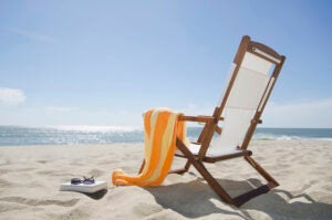 An orange striped towel rests on the arm of a wooden beach chair that's on the sand facing the ocean. A book and sunglasses on the sand next to the chair.