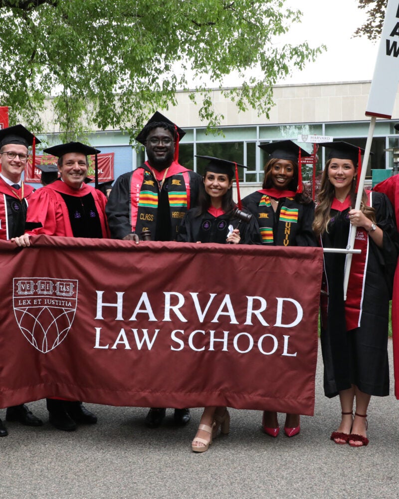 Class Marshals in commencement regalia holding a banner that says Harvard Law School pose for a group shot with Dean Manning and Dean of Students Stephen Ball