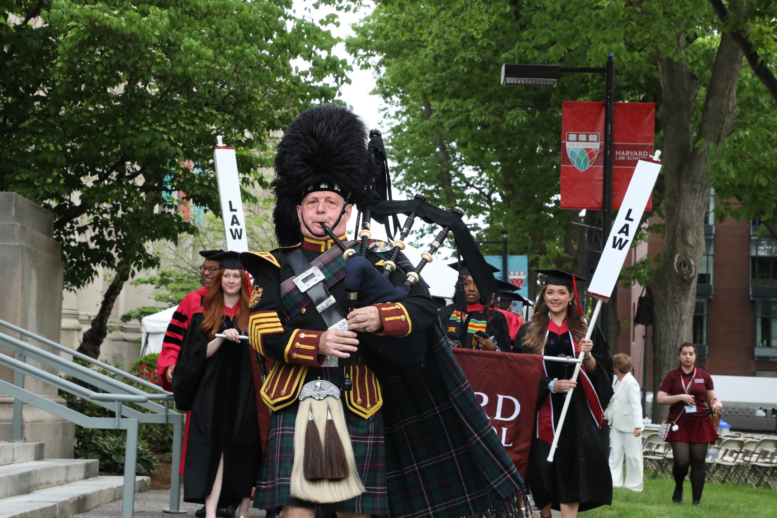 A bagpiper leads students in commencement regalia along a sidewalk on the HLS campus