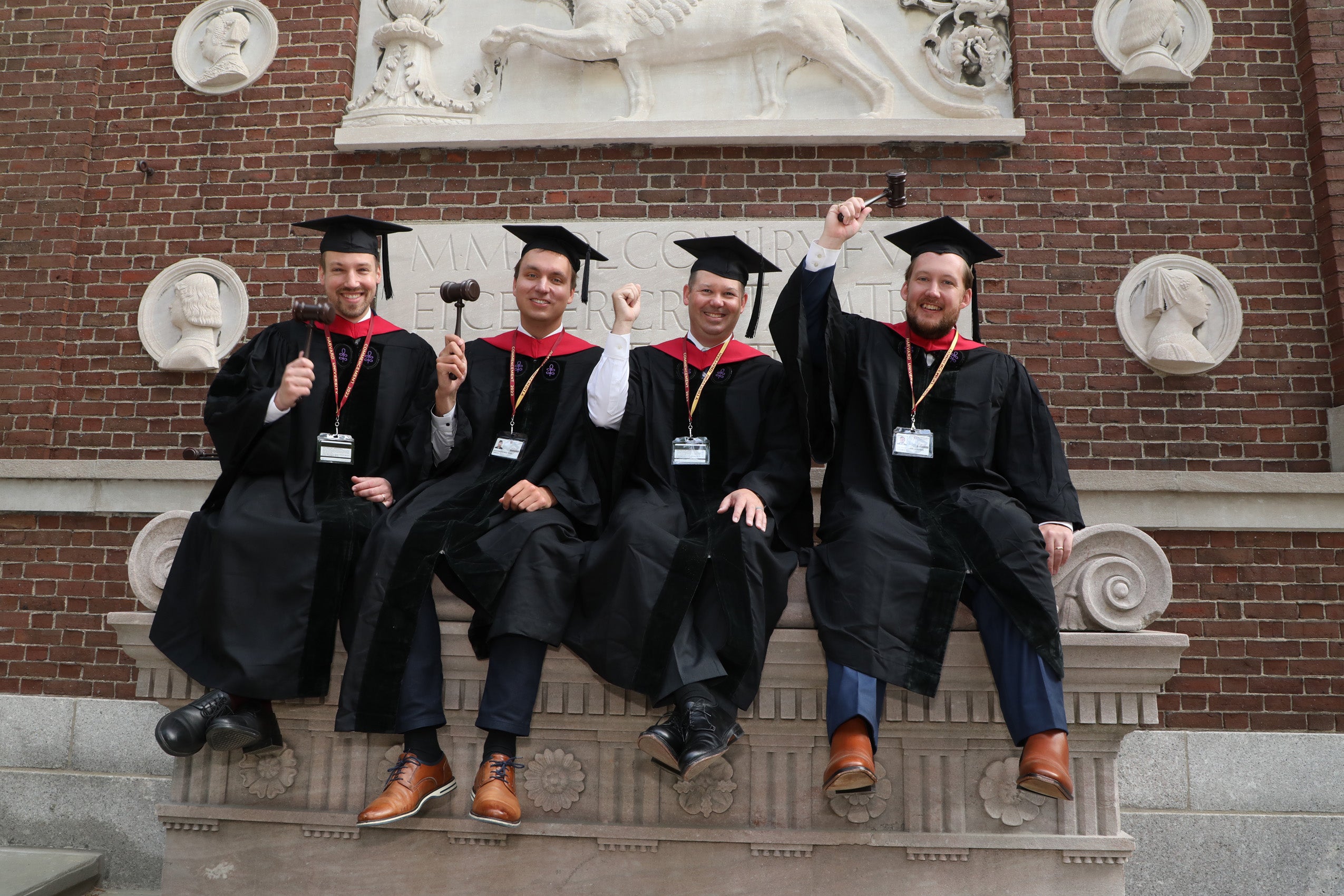 A group of graduates in commencement regalia sit on an stone bench and hold up their gavels