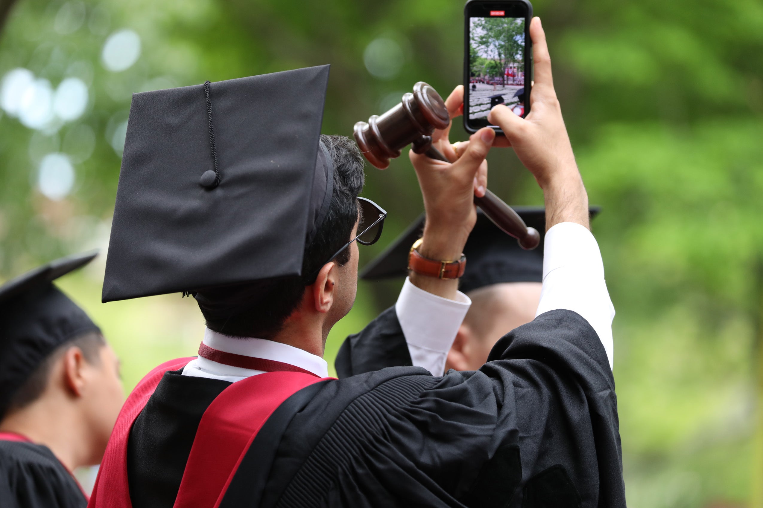 A man wearing a cap and gown holding a cell phone captures Harvard Yard on his screen
