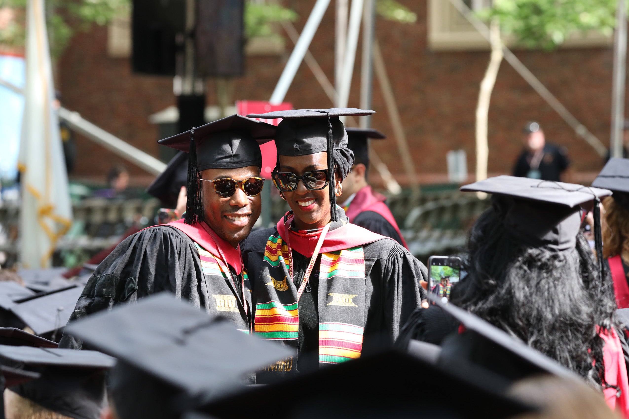 Several students in caps and gowns stand up and wave and gesture in Harvard Yard