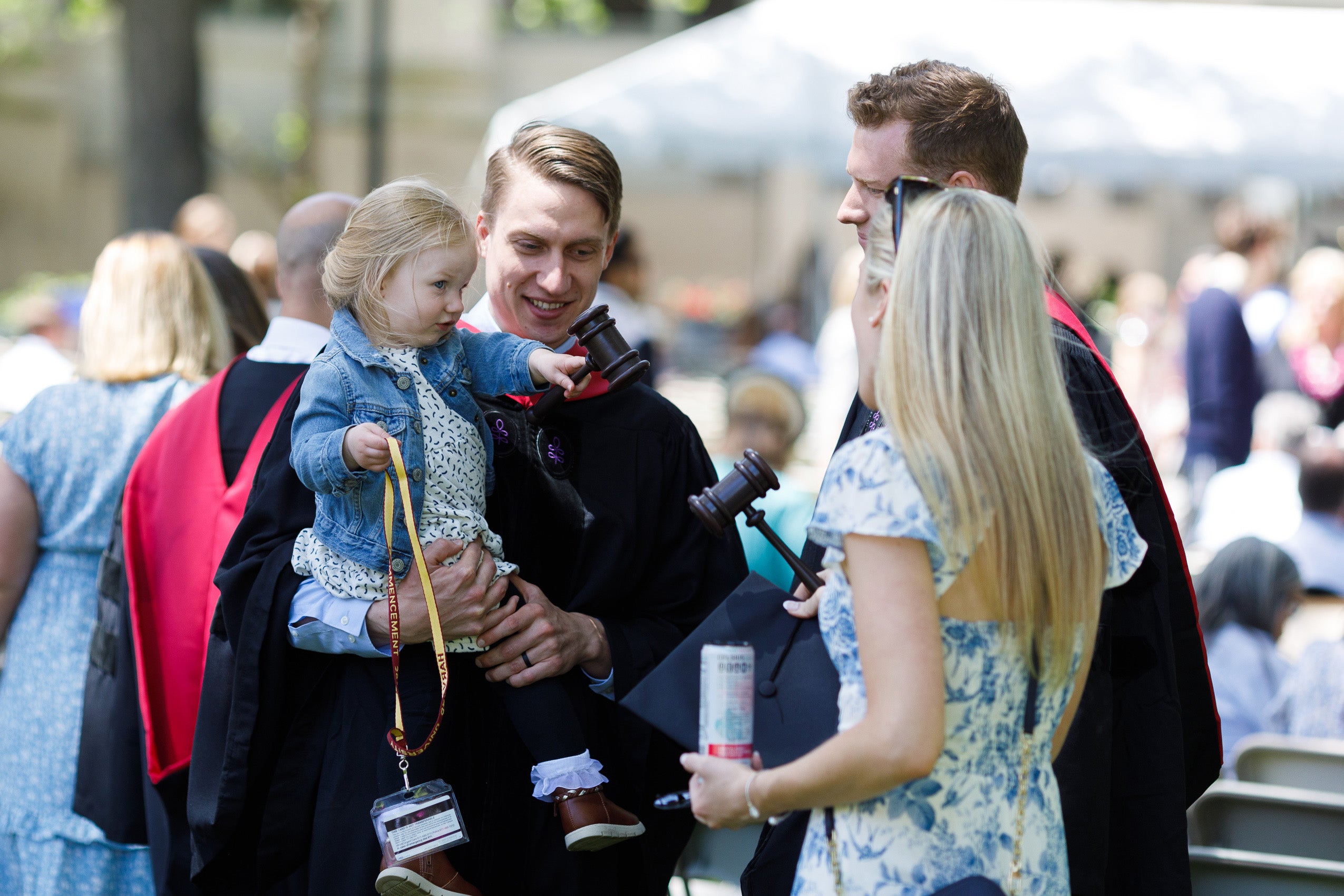 A man in cap and gown talks to a couple one of whom is also a graduate as he holds a young girl who has his gavel in her hand