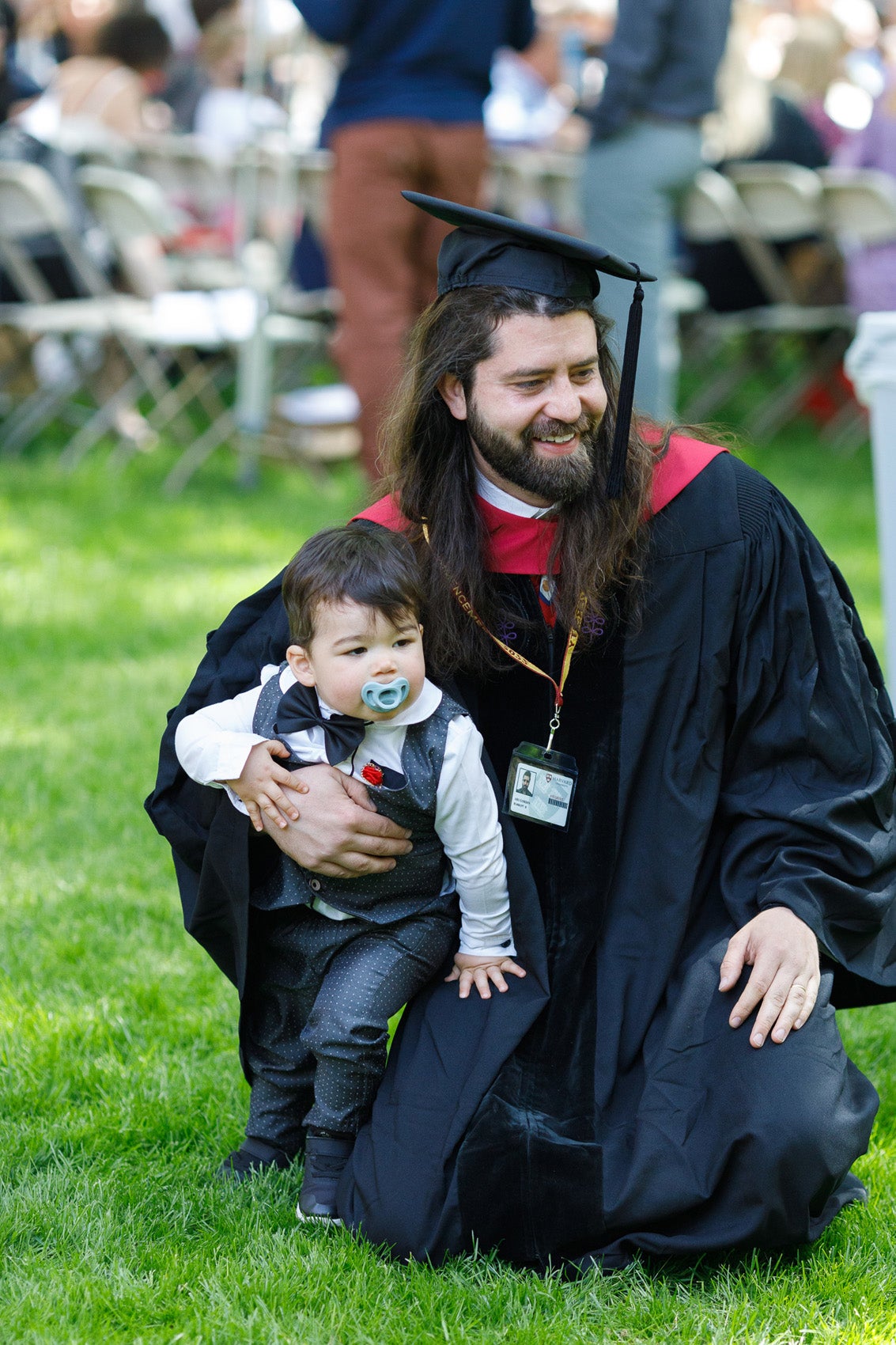 A man wearing a cap and gown bends on the green grass with a toddler wearing a bow ties and dress pants and white shirt
