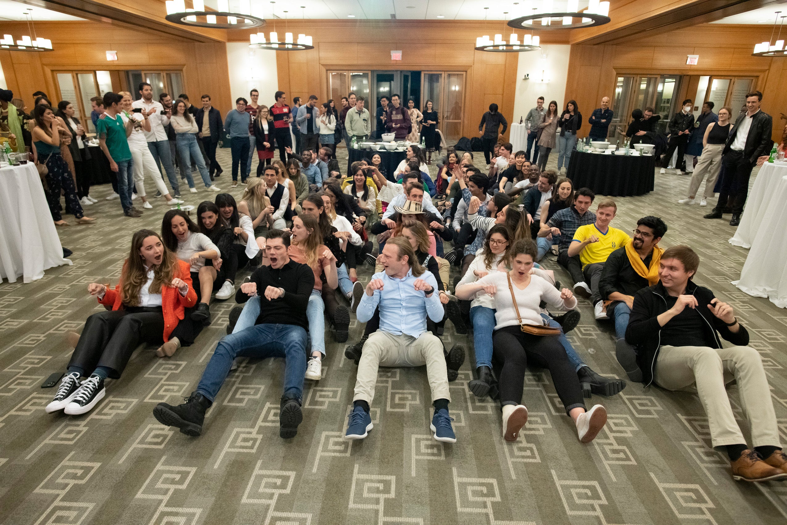 Rows of students at an event sit on the ground and pretend to row together