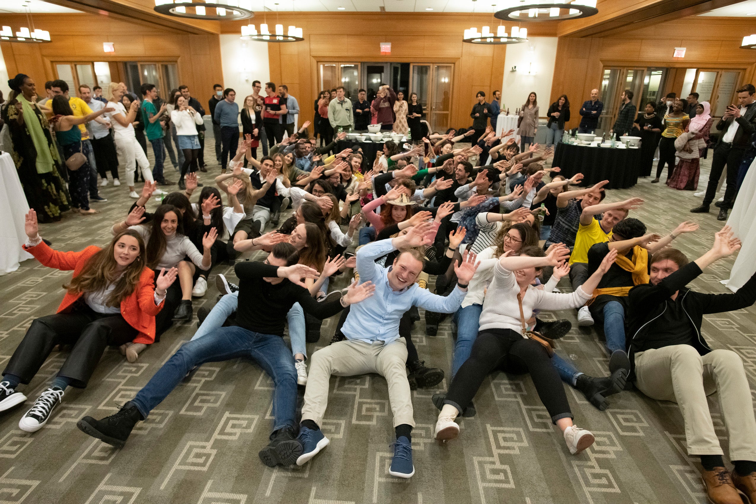 Rows of students at an event sit on the ground wave their hands to the side overhead as they pretend to row a boat together