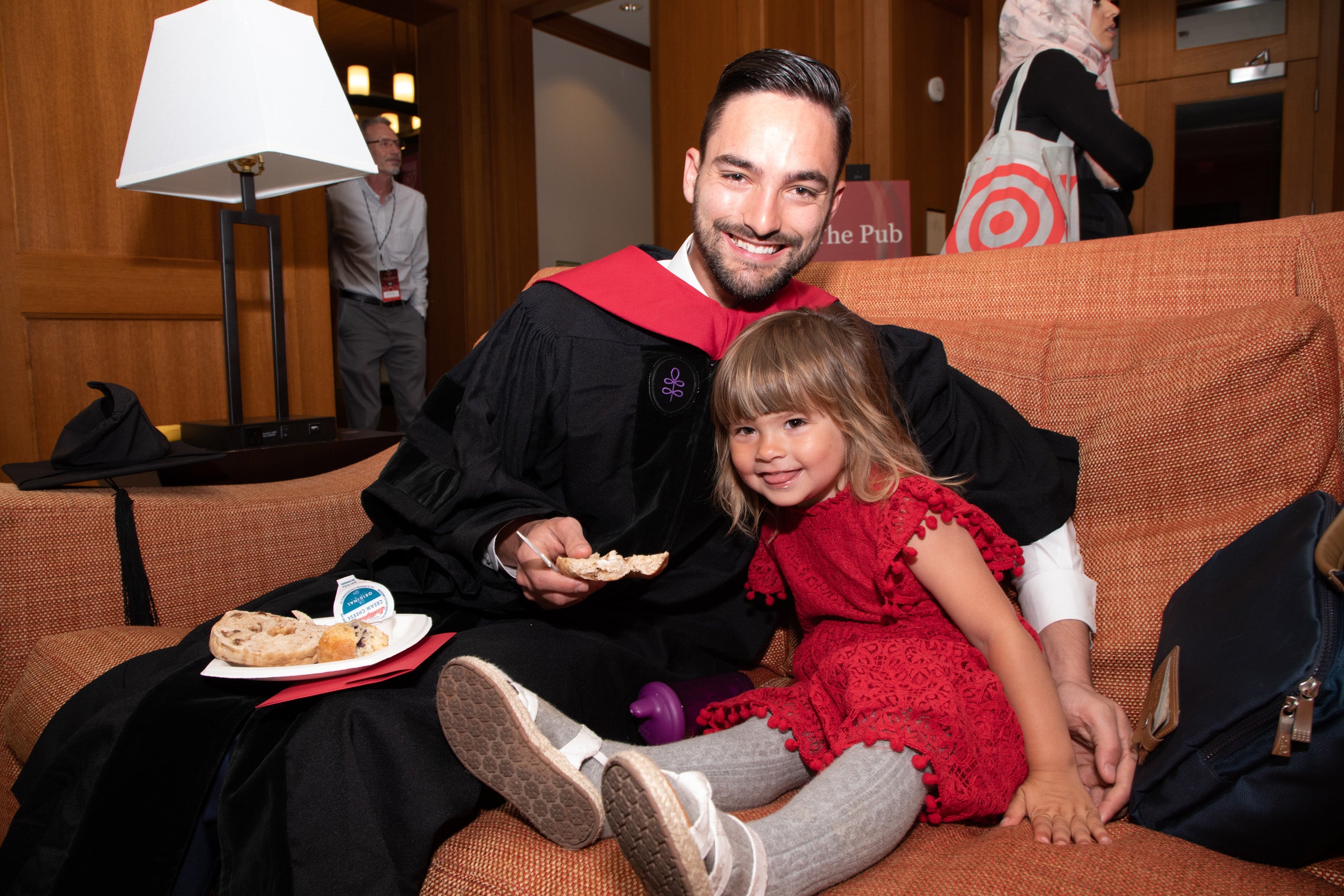 A man in a cap and gown sits next to a young girl on a couch located inside an HLS building