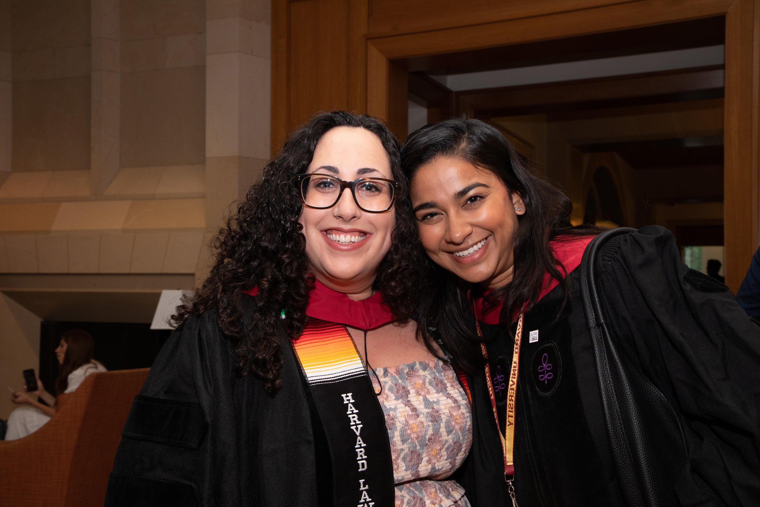Two women in graduation gowns pose inside an HLS building for a photo