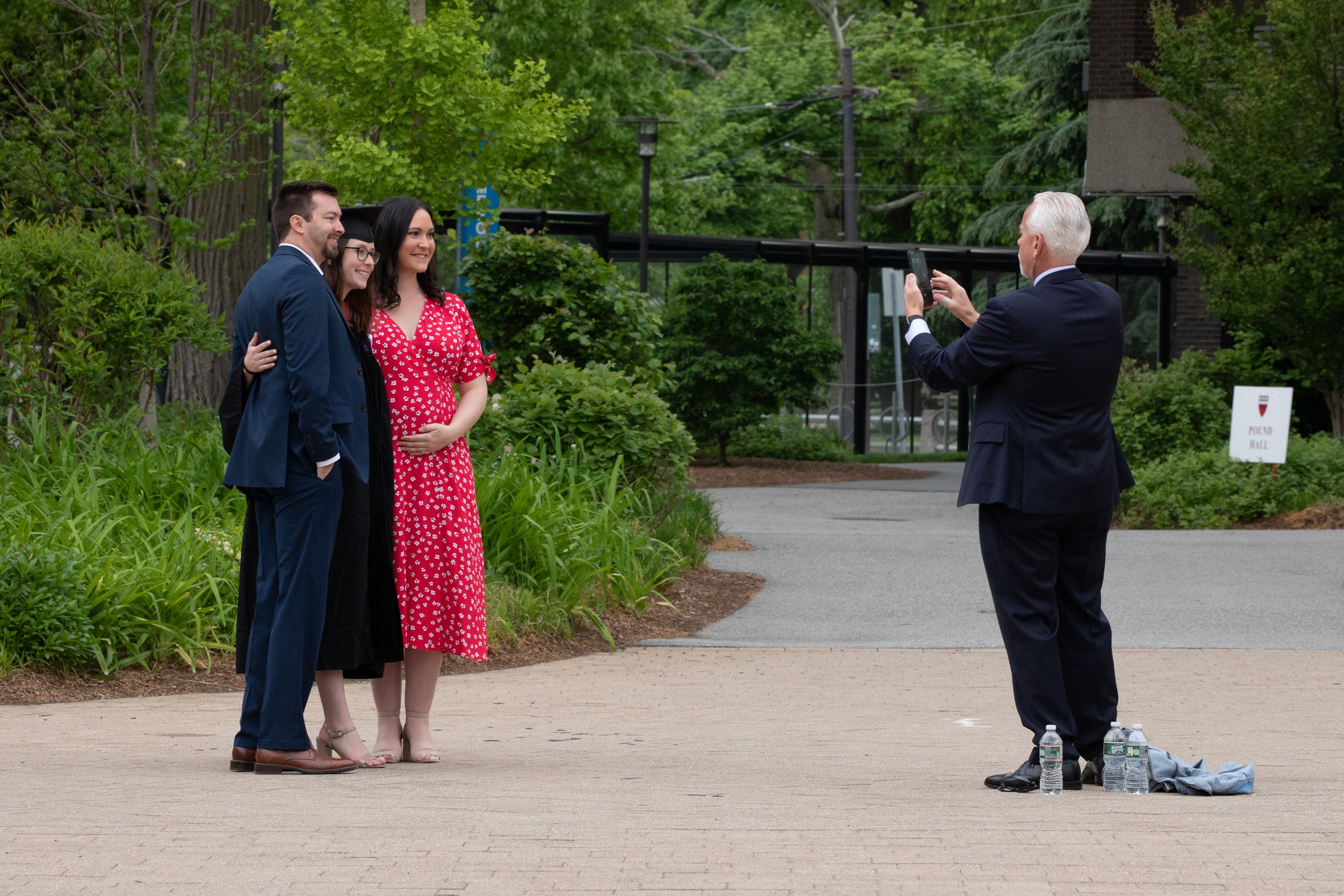 A man takes a photo of a graduate wearing a cap and gown standing between a woman in a red and white dress and a man in a blue suit