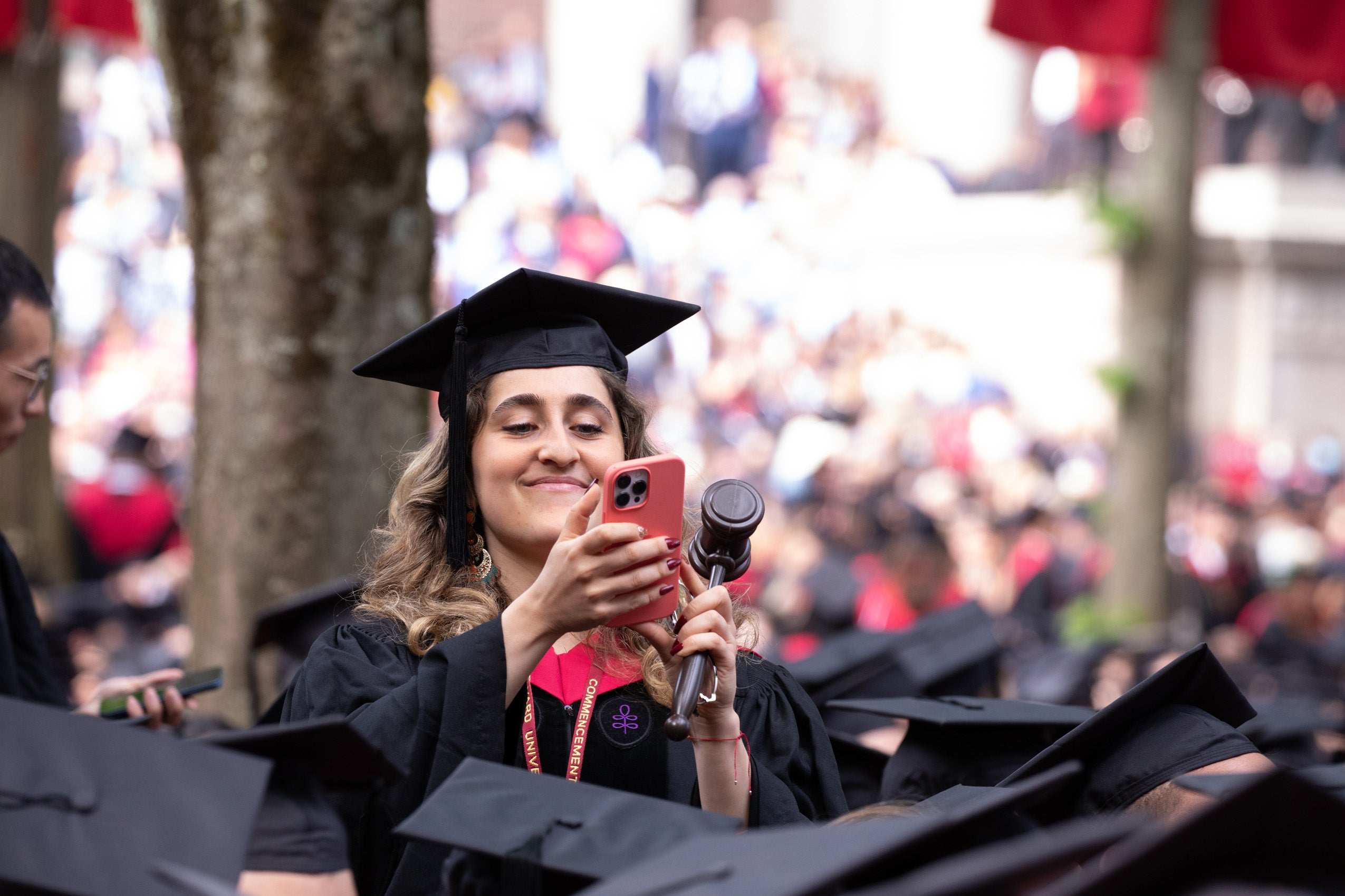 A graduate stands up above a sea of graduate caps and smiles at something on her cell phone