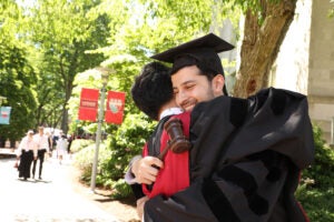 Two graduates wearing caps and gowns hug on a pathway on campus