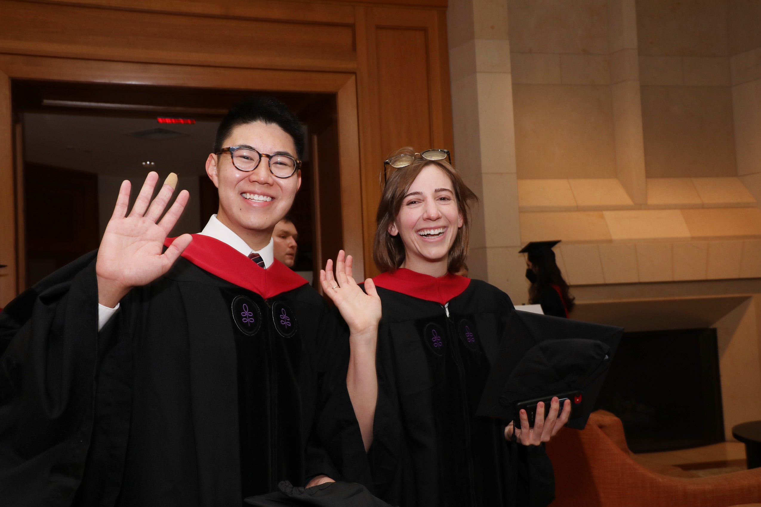 A man and a woman wearing commencement robes wave at the camera