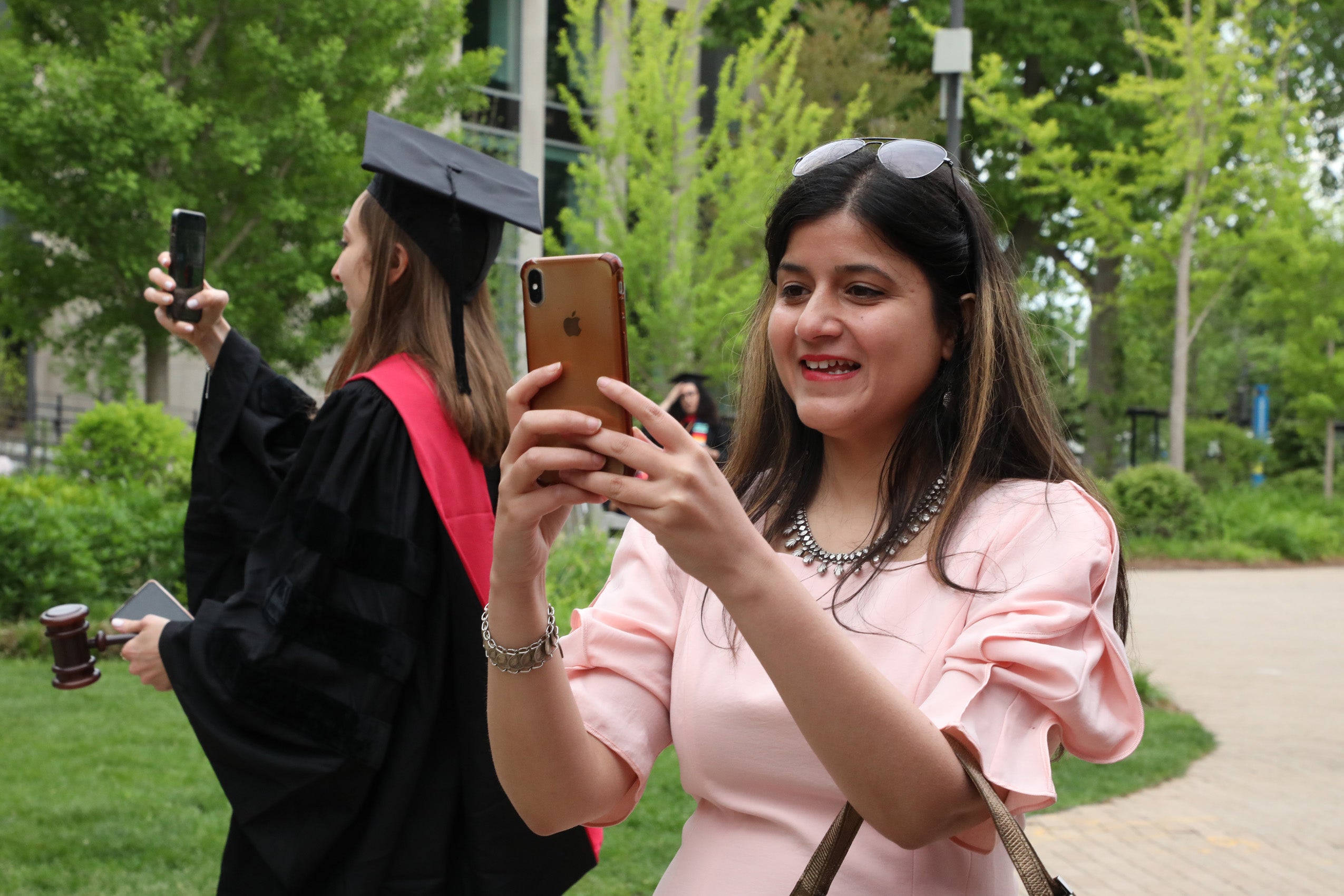 A woman wearing commencement regalia and a gavel and a woman in a pink dress both holding cel phones standing back to back