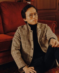 A man in a light brown plaid jacket sits in front of a rusty red couch