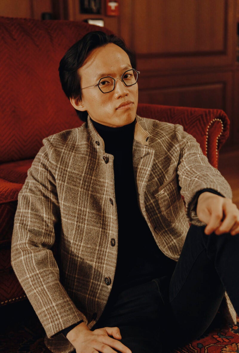 A man in a light brown plaid jacket over a black turtleneck sweater sits in front of a rusty red couch